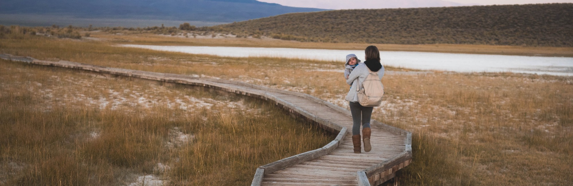 woman walks on boardwalk next to lake and brown grass while carrying baby